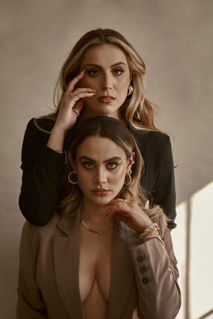Clarisa and Valeria two women with striking features pose elegantly against a muted backdrop. Clarisa with her hand on her face exudes a contemplative aura while Valeria's direct gaze captures attention. Both are adorned with jewelry adding to the sophistication of the scene.